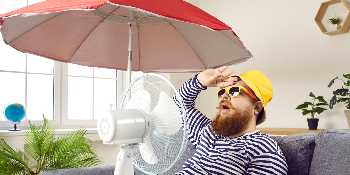 A man sits indoors under a sun umbrella and in front of a fan, exasperated by the heat. 